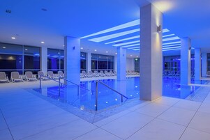 Un área de piscina interior moderna con barandas de cristal transparente, sillas de descanso e iluminación ambiental azul.