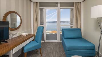 Ett hotellrum med ett skrivbord i trä, en tv, två blå fåtöljer, en soffa och utsikt över havet genom en glasdörr.