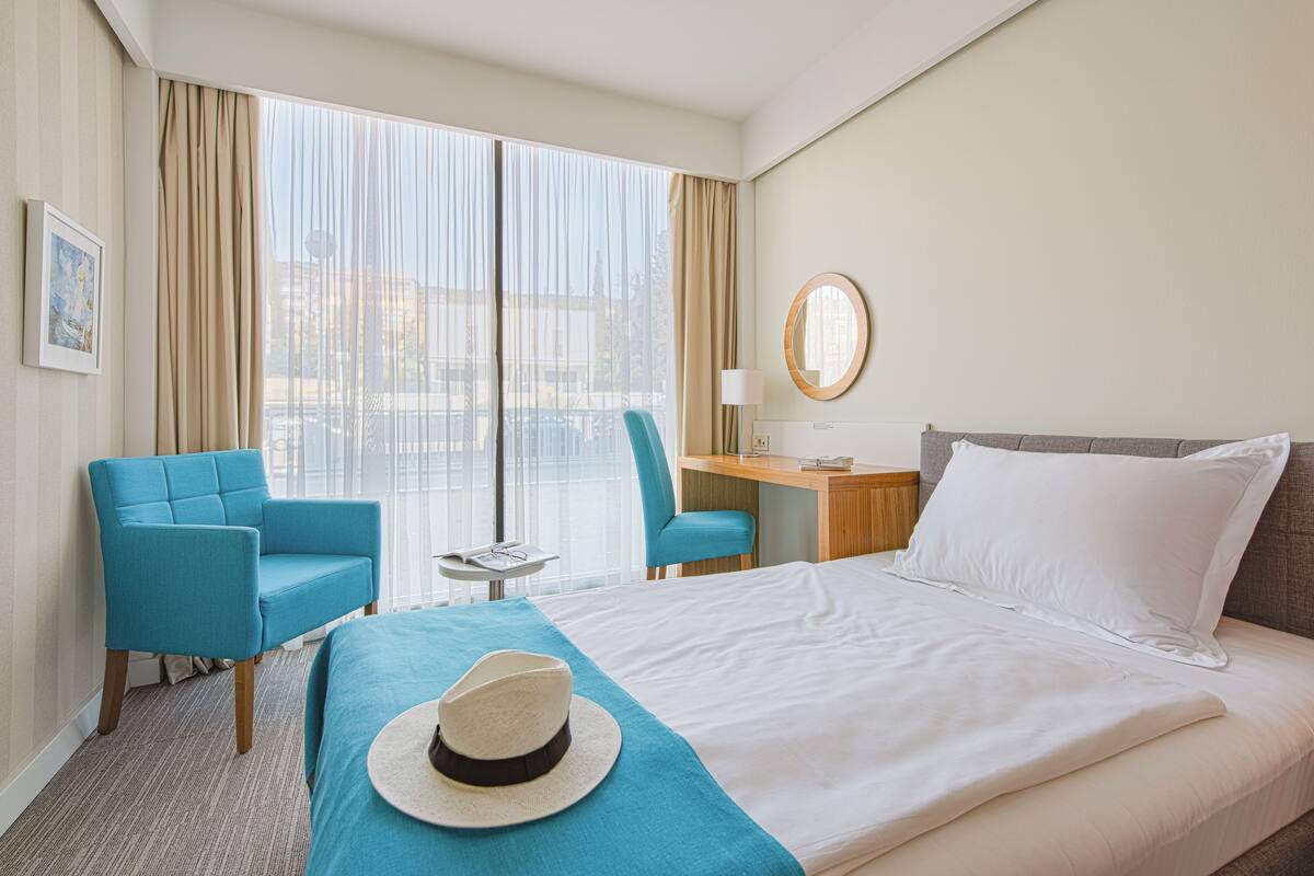 A hotel room with a bed, two chairs, a desk, a lamp, and a view of the city through large windows.