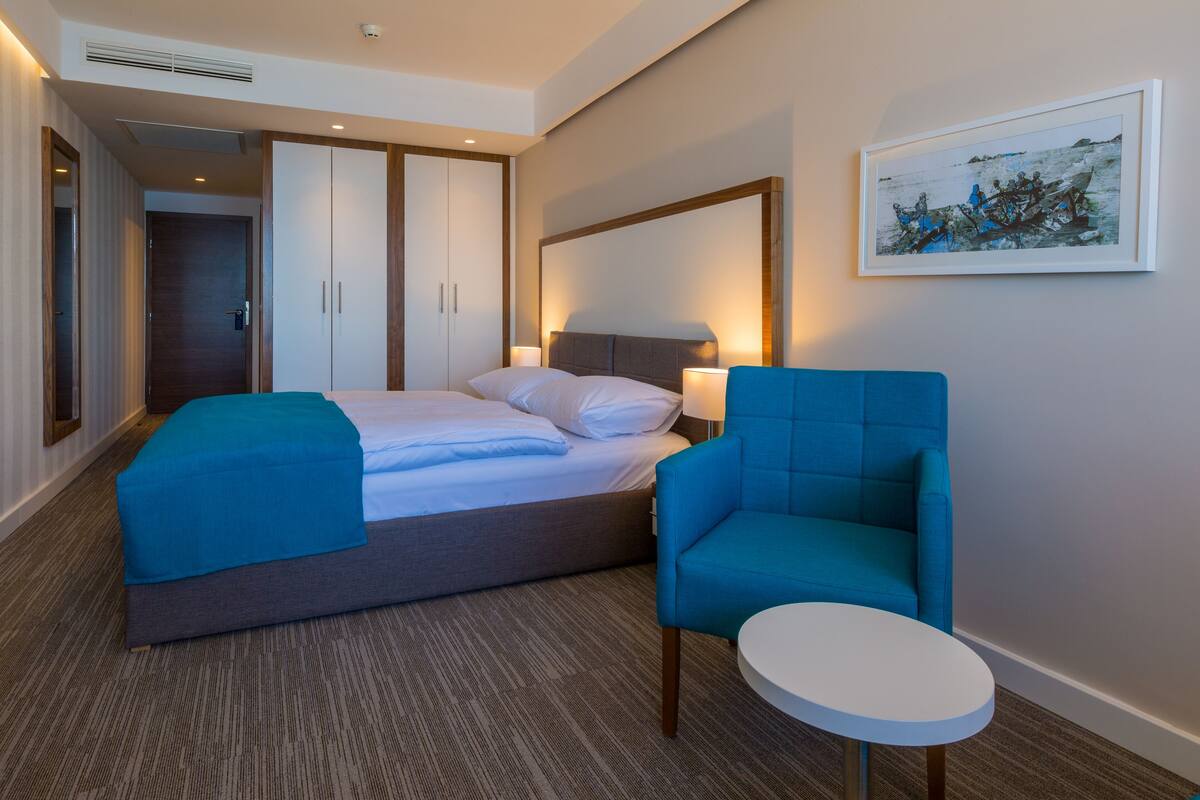 A hotel room with a bed, a blue armchair, a small round table, and a framed picture on the wall.