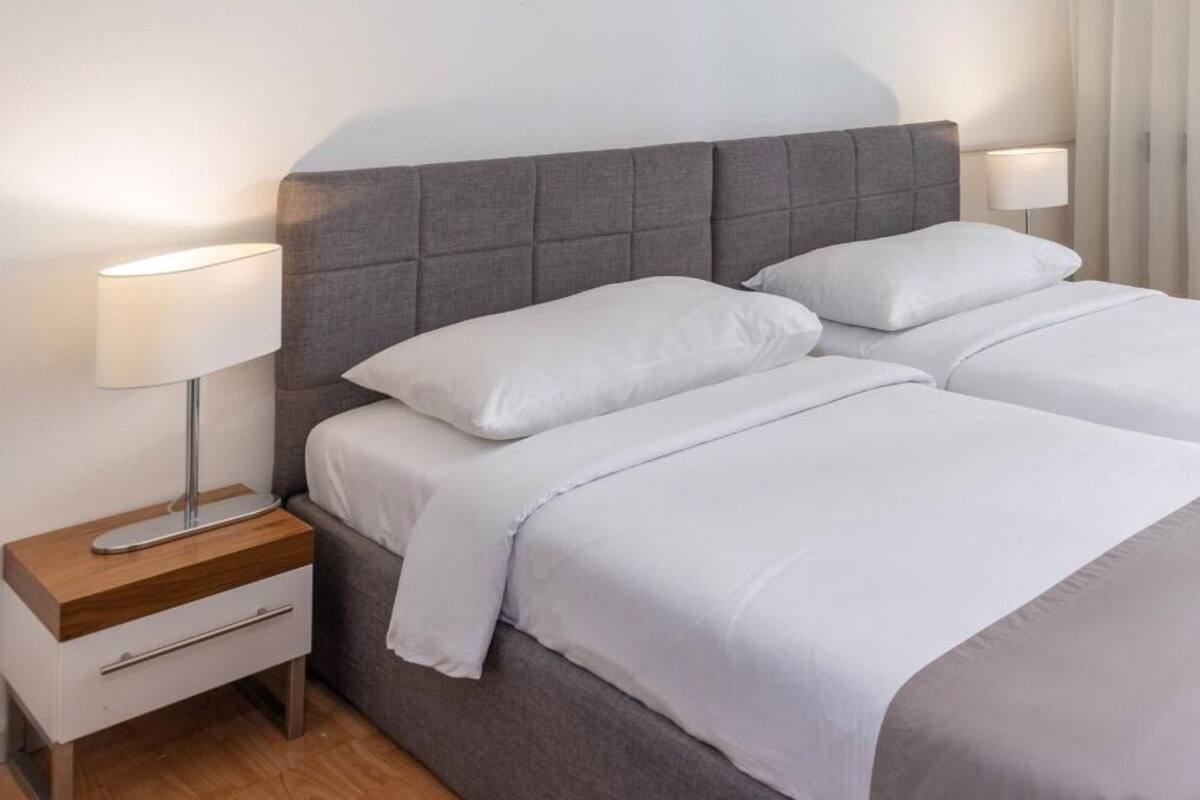 A double bed with grey headboard and white bedding, a bedside table with a lamp, and a window with curtains.