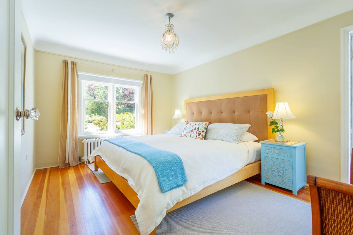 A bedroom with a large bed, a nightstand with a lamp, a window with curtains, and a radiator.