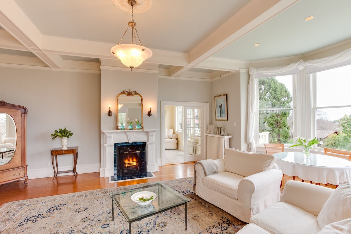 A spacious living room with a fireplace, white sofas, a glass coffee table, and a large window with curtains.
