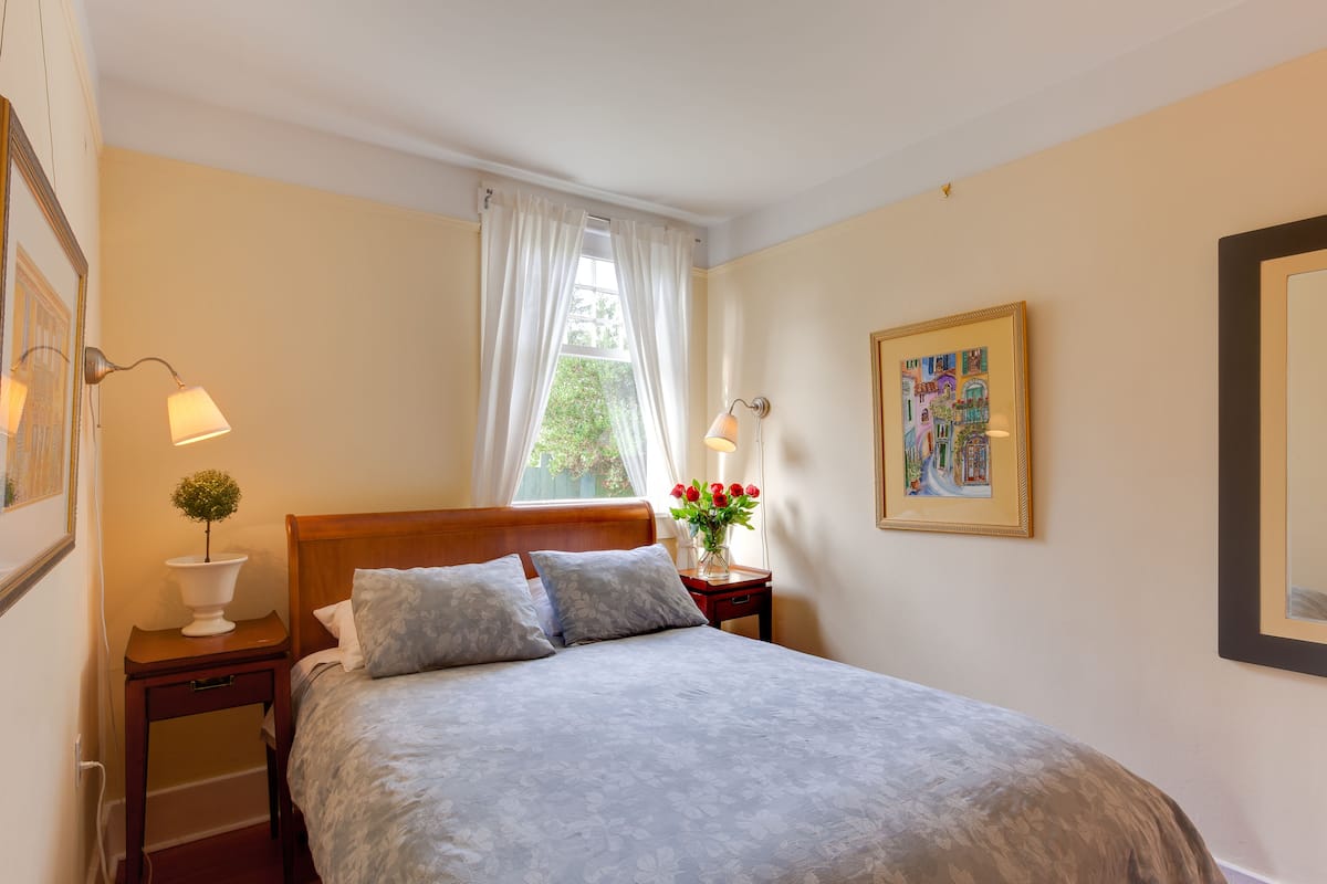 A neatly made bed with a floral bedspread, a bedside table with a lamp, a vase with flowers, and a framed picture on the wall.