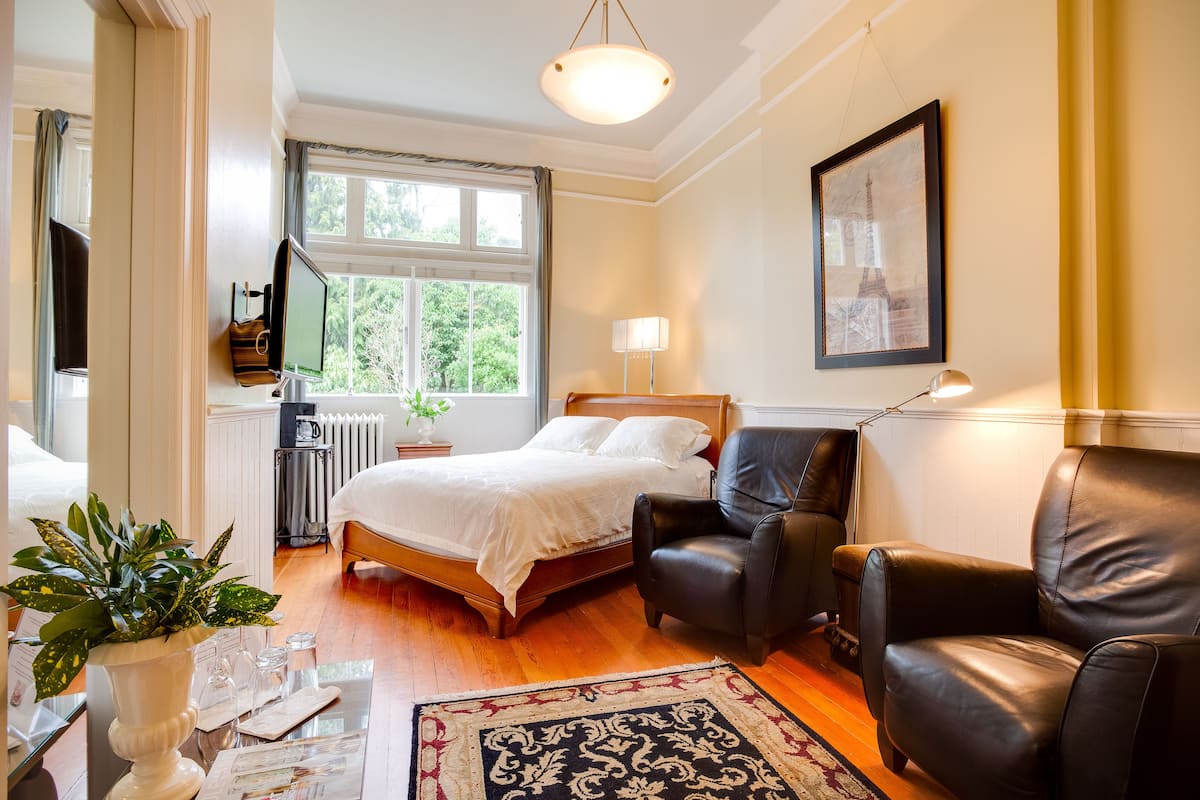 A well-lit bedroom with a bed, two chairs, a desk with a lamp, a window with curtains, and a framed picture on the wall.