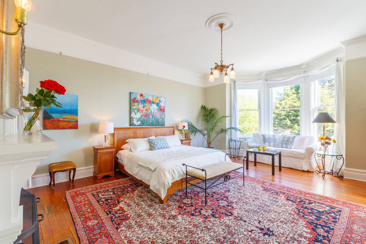 A bedroom with a large bed, a sitting area with a sofa, a dining table, and a view of the outdoors through large windows.