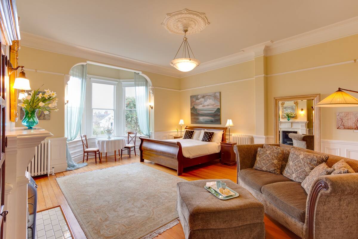 A well-lit bedroom with a large bed, a sitting area with a sofa and armchair, a dining table, and a view of the outdoors through a window.