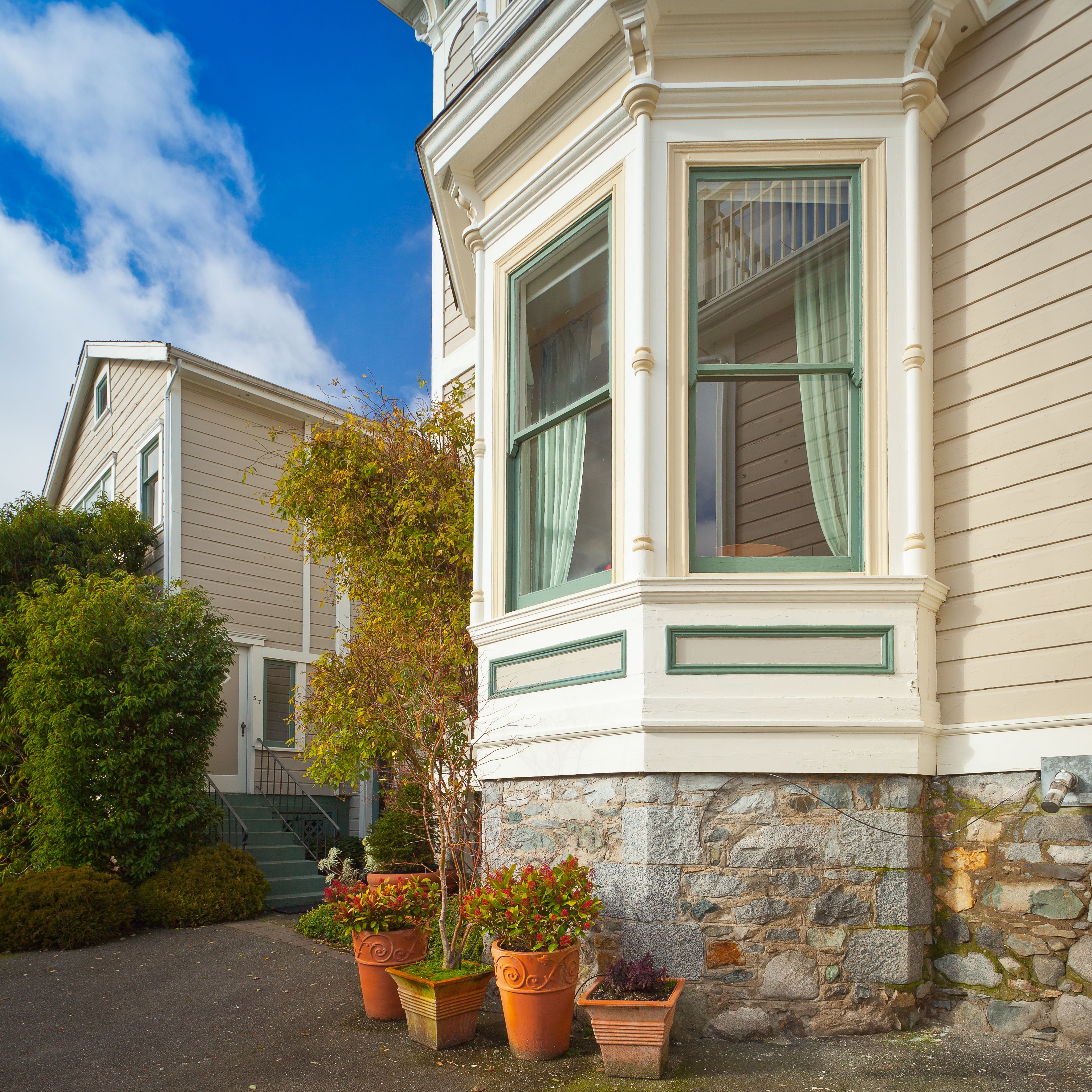 A corner of a building with a stone foundation, a bay window with green trim, and several potted plants.