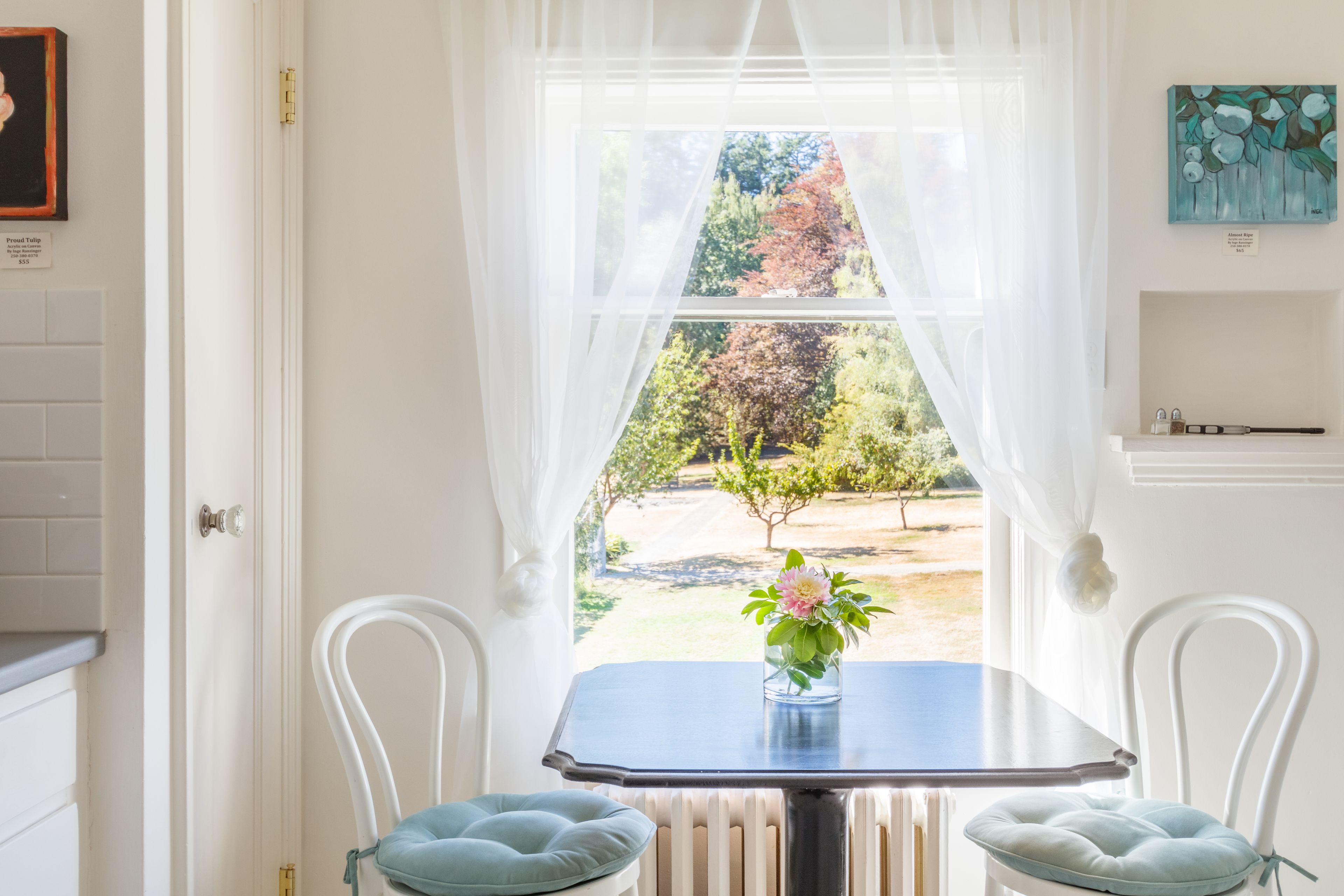A room with a table and two chairs, a window with sheer curtains, and a view of a garden.