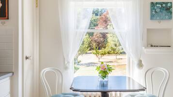 A room with a table and two chairs, a window with sheer curtains, and a view of a garden.