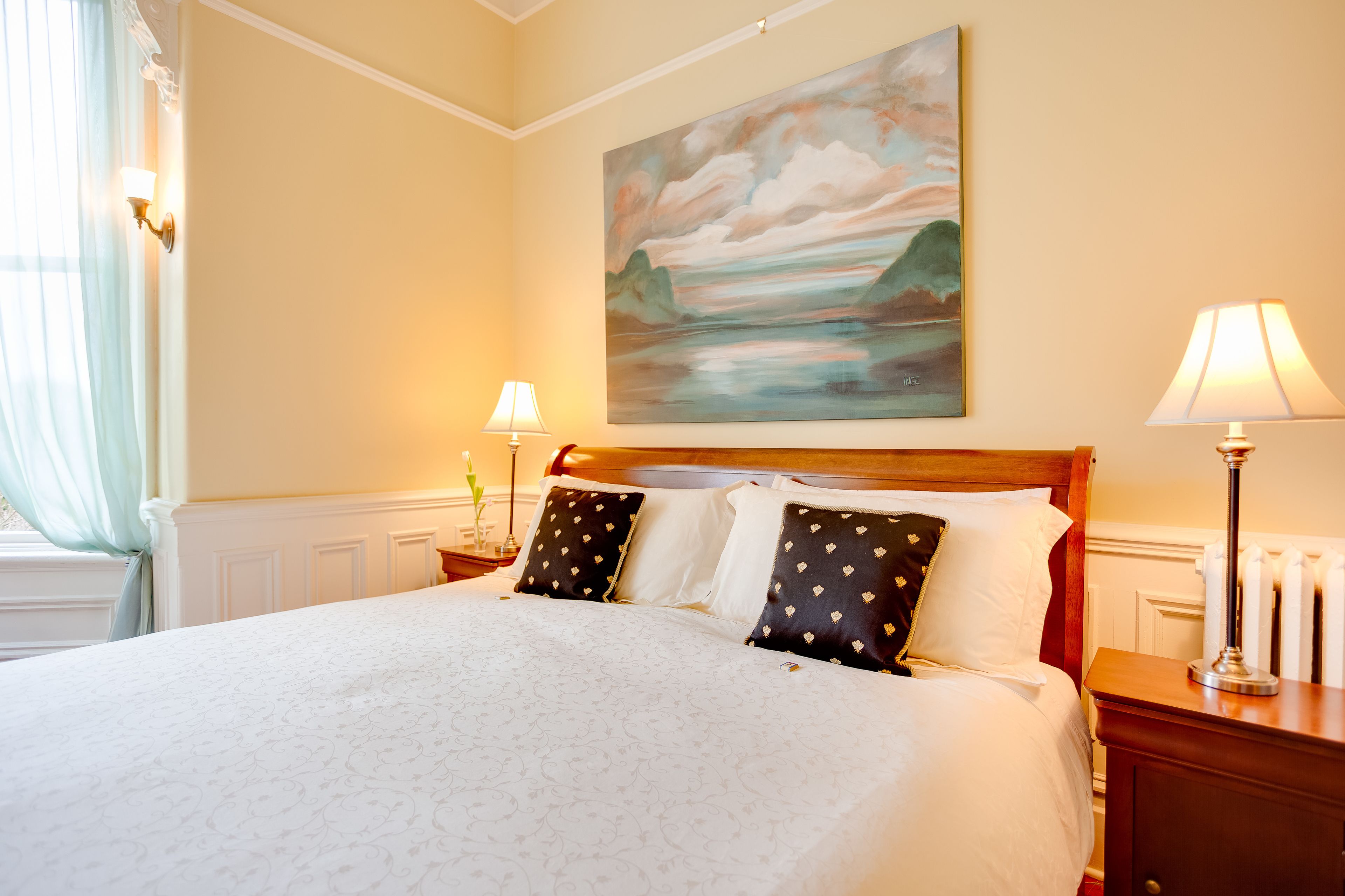 A neatly made bed with a white duvet and patterned bedding, a painting of a landscape on the wall, and two bedside lamps on wooden nightstands.