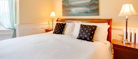 A neatly made bed with a white duvet and patterned bedding, a painting of a landscape on the wall, and two bedside lamps on wooden nightstands.