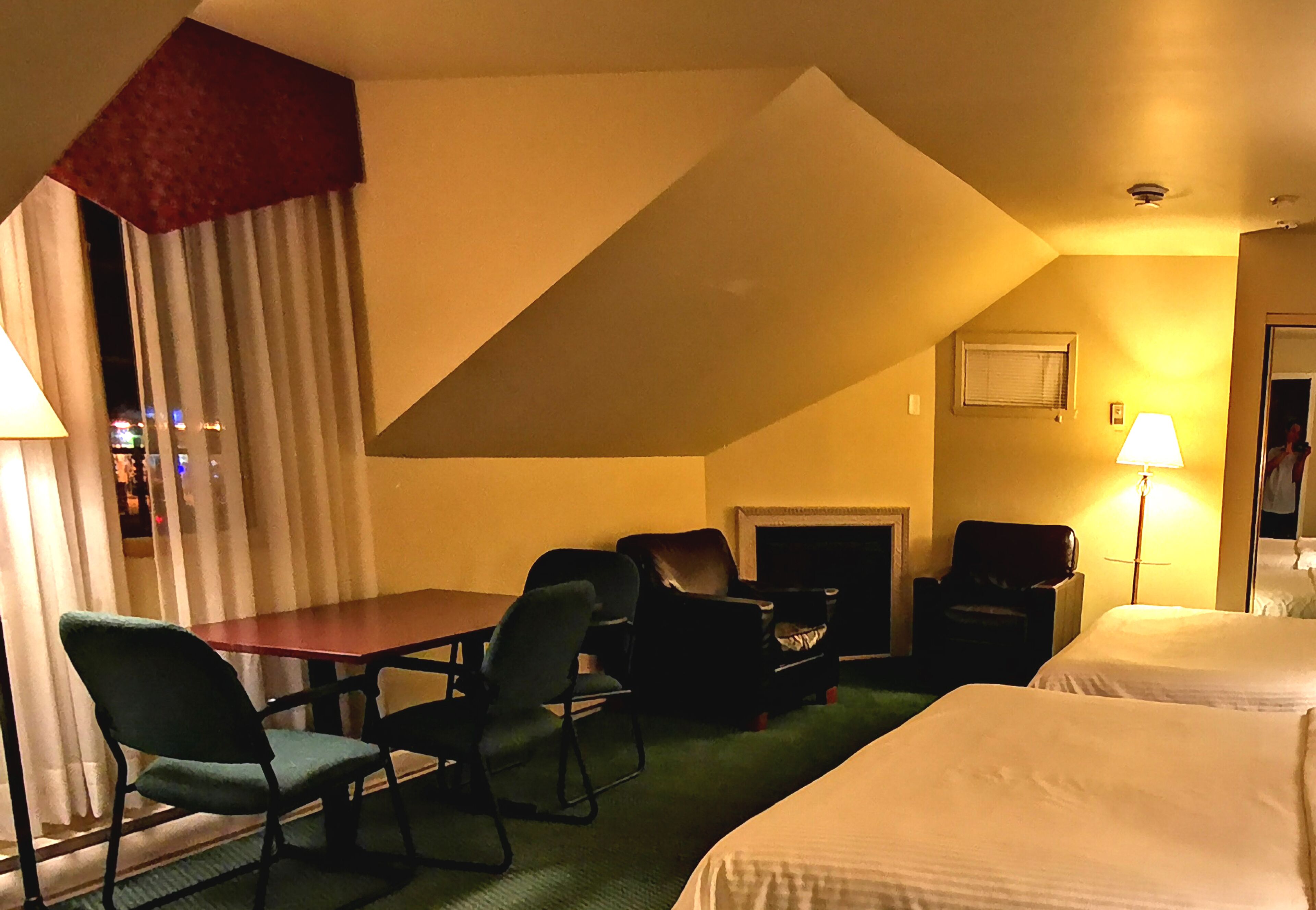 A hotel room with a bed, a desk, two chairs, a fireplace, and a lamp.
