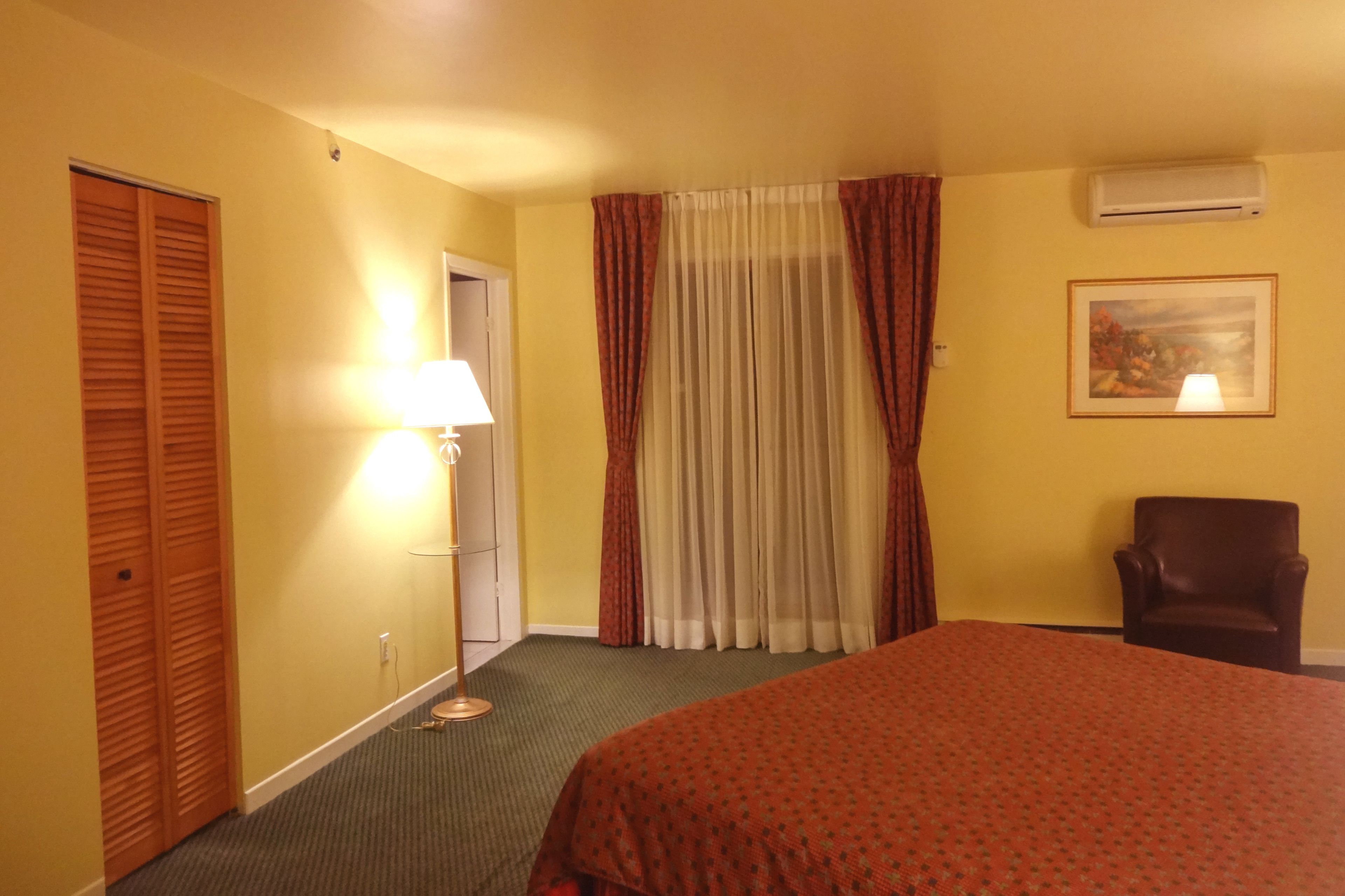 Habitación de hotel con cama, silla, lámpara, un cuadro y unidad de aire acondicionado.