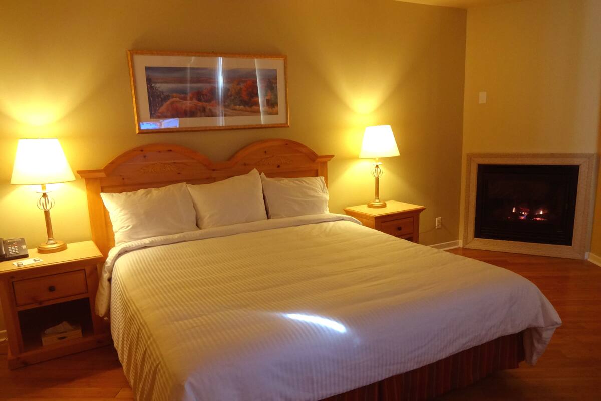 A neatly made bed with a wooden headboard, two bedside lamps, a nightstand with a phone, and a fireplace in the corner.