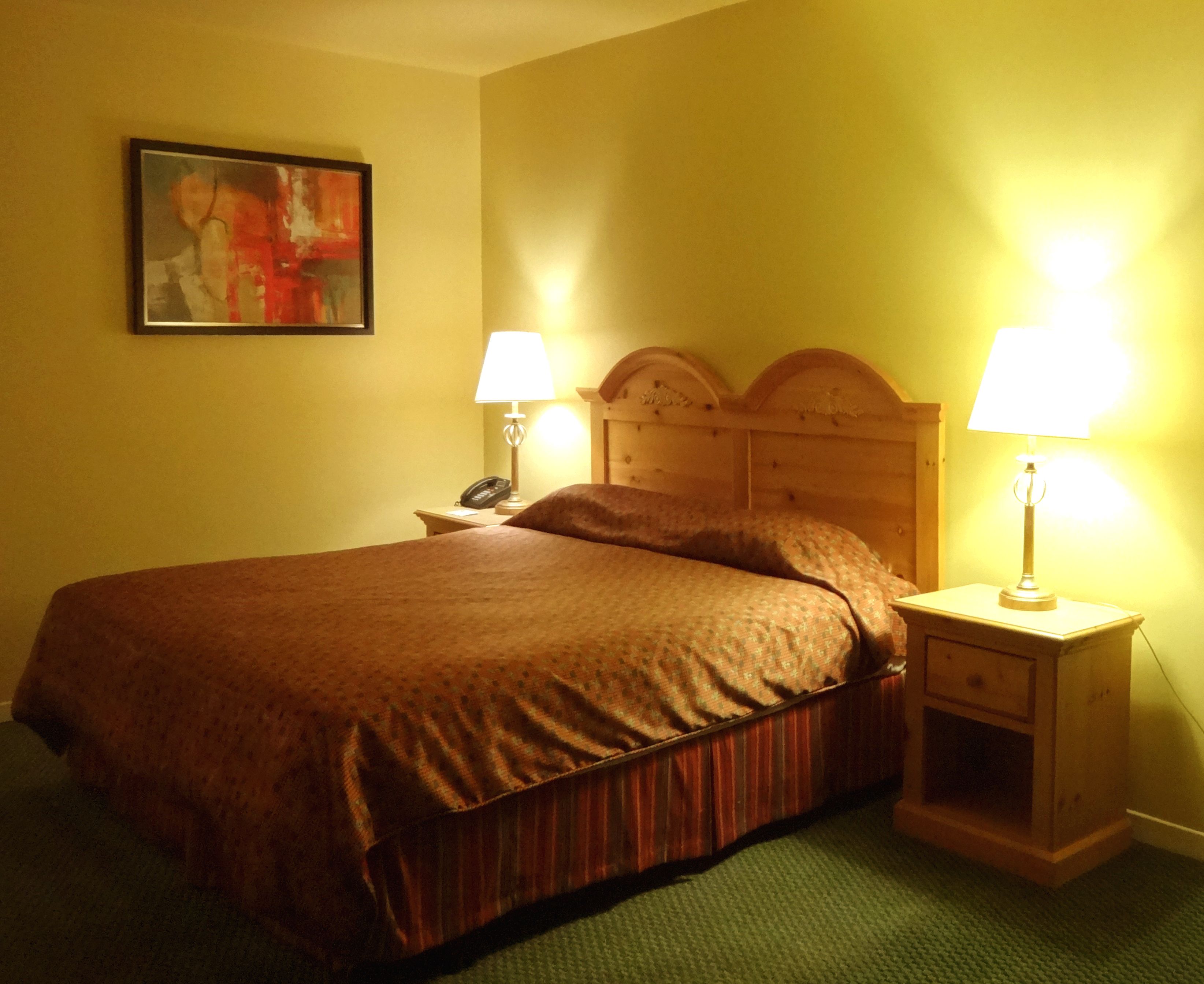 A hotel room with a wooden headboard, a bed with a brown bedspread, two bedside lamps, a telephone, and a painting on the wall.