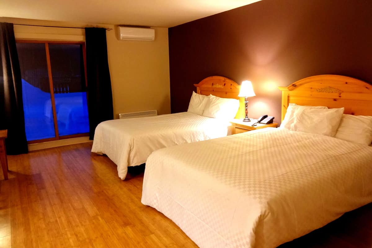 A hotel room with two beds, a nightstand with a lamp, a window with curtains, and a sliding door.