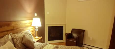A hotel room with a bed, a nightstand with a lamp, a fireplace, and a leather chair.
