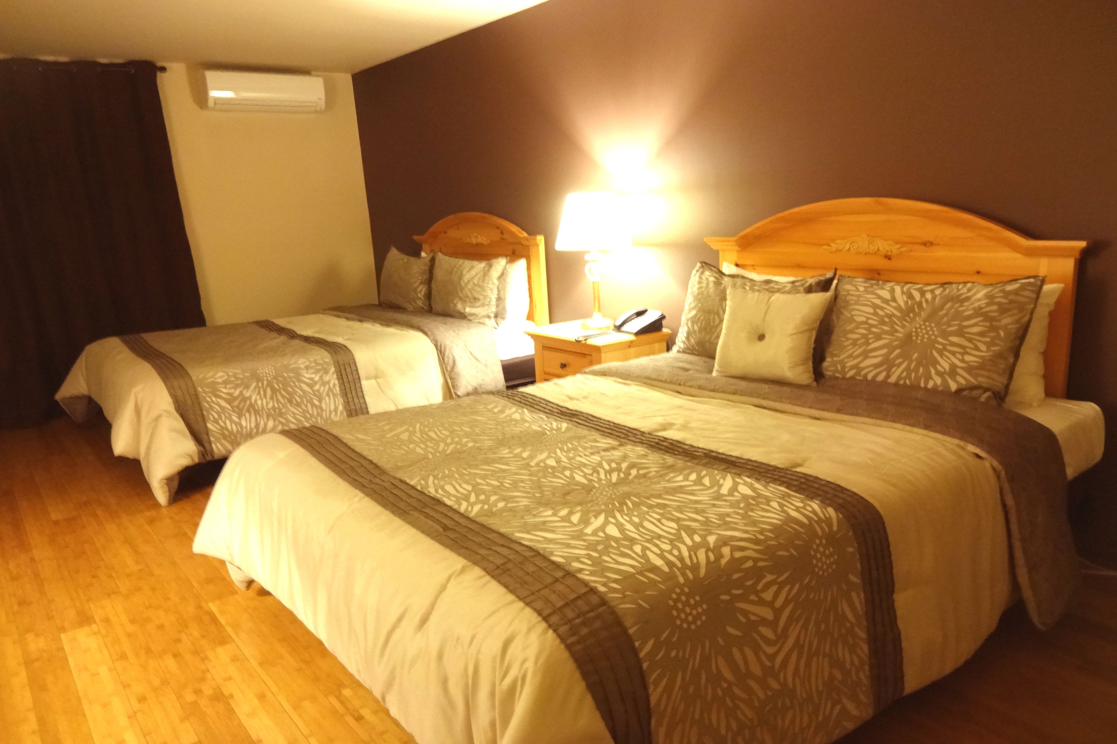 A hotel room with two beds, a wooden headboard, a bedside table with a lamp, and a wall-mounted air conditioner.