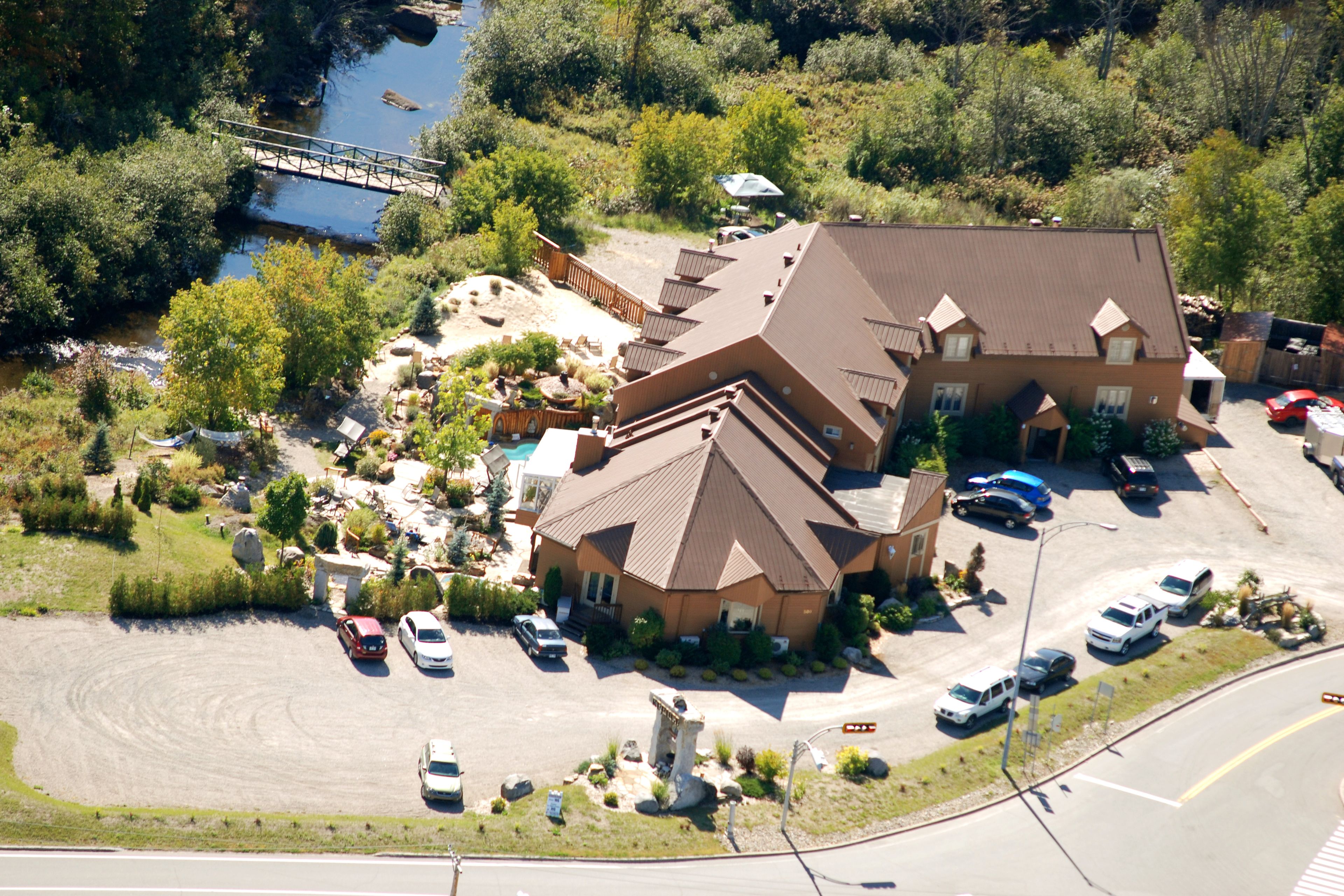 An aerial view of a large house with a steep roof, surrounded by a parking lot and a small bridge over a stream.