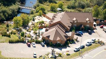 An aerial view of a large house with a steep roof, surrounded by a parking lot and a small bridge over a stream.
