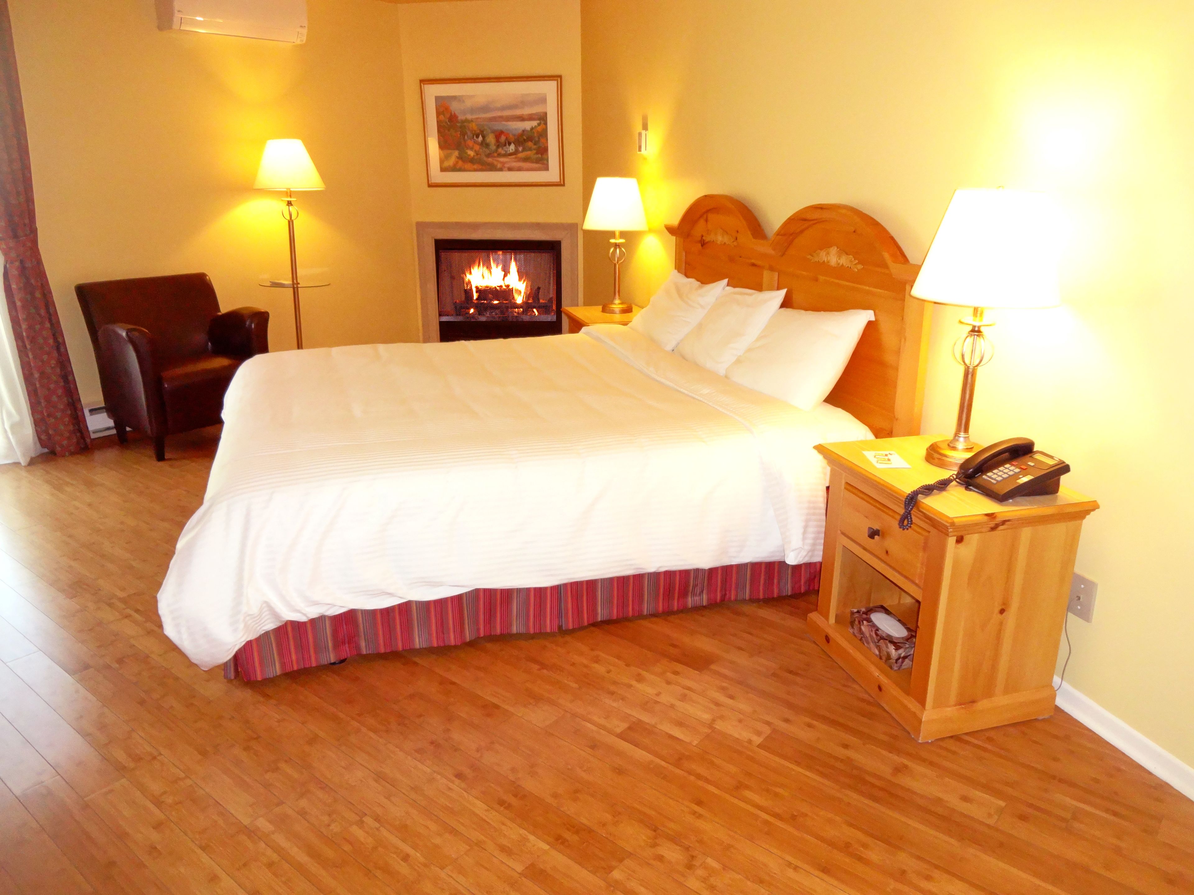 A neatly made bed with white linens, a wooden nightstand with a telephone, and a fireplace in the background.