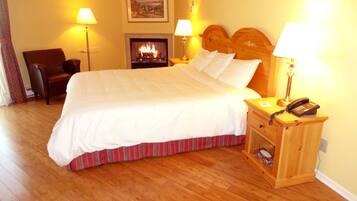 A neatly made bed with white linens, a wooden nightstand with a telephone, and a fireplace in the background.