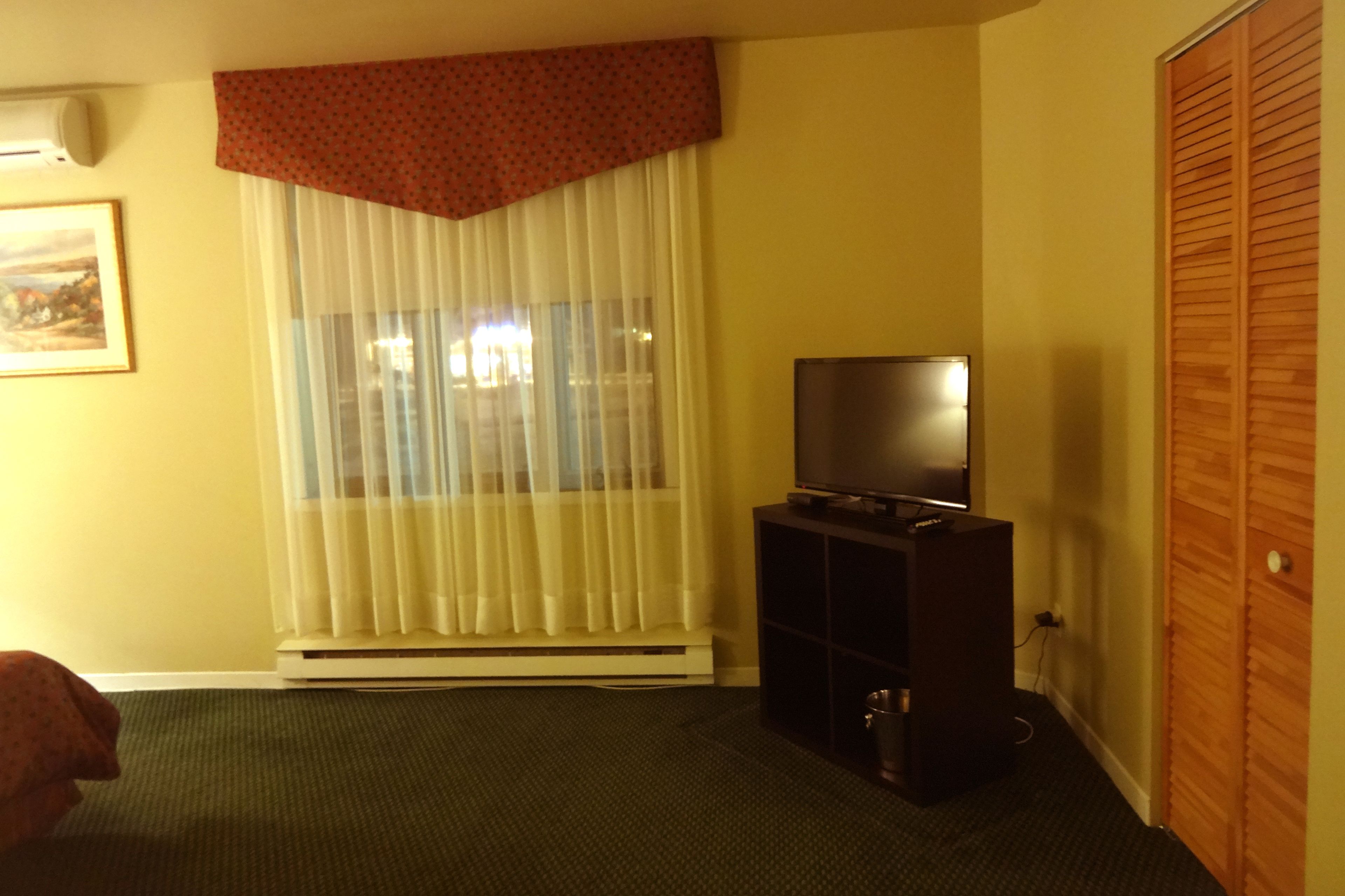 Ein Zimmer mit Fernseher auf einem Standfuß, einem Heizkörper, einem Fenster mit durchscheinenden Vorhängen und einer Holztür.