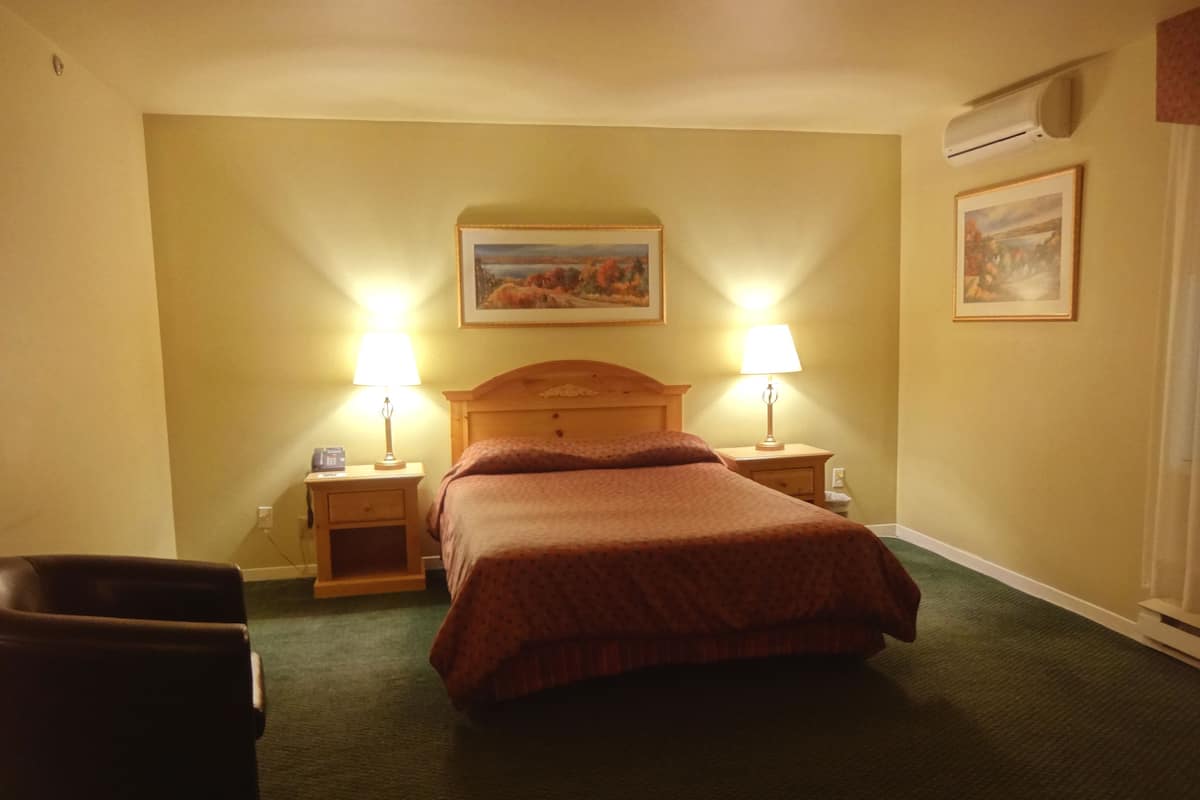 A hotel room with a bed, two bedside lamps, a wooden headboard, a nightstand, a chair, and a painting on the wall.