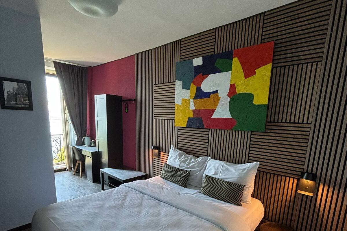 A modern hotel room with a large bed, a desk, and a colorful abstract painting on the wall.