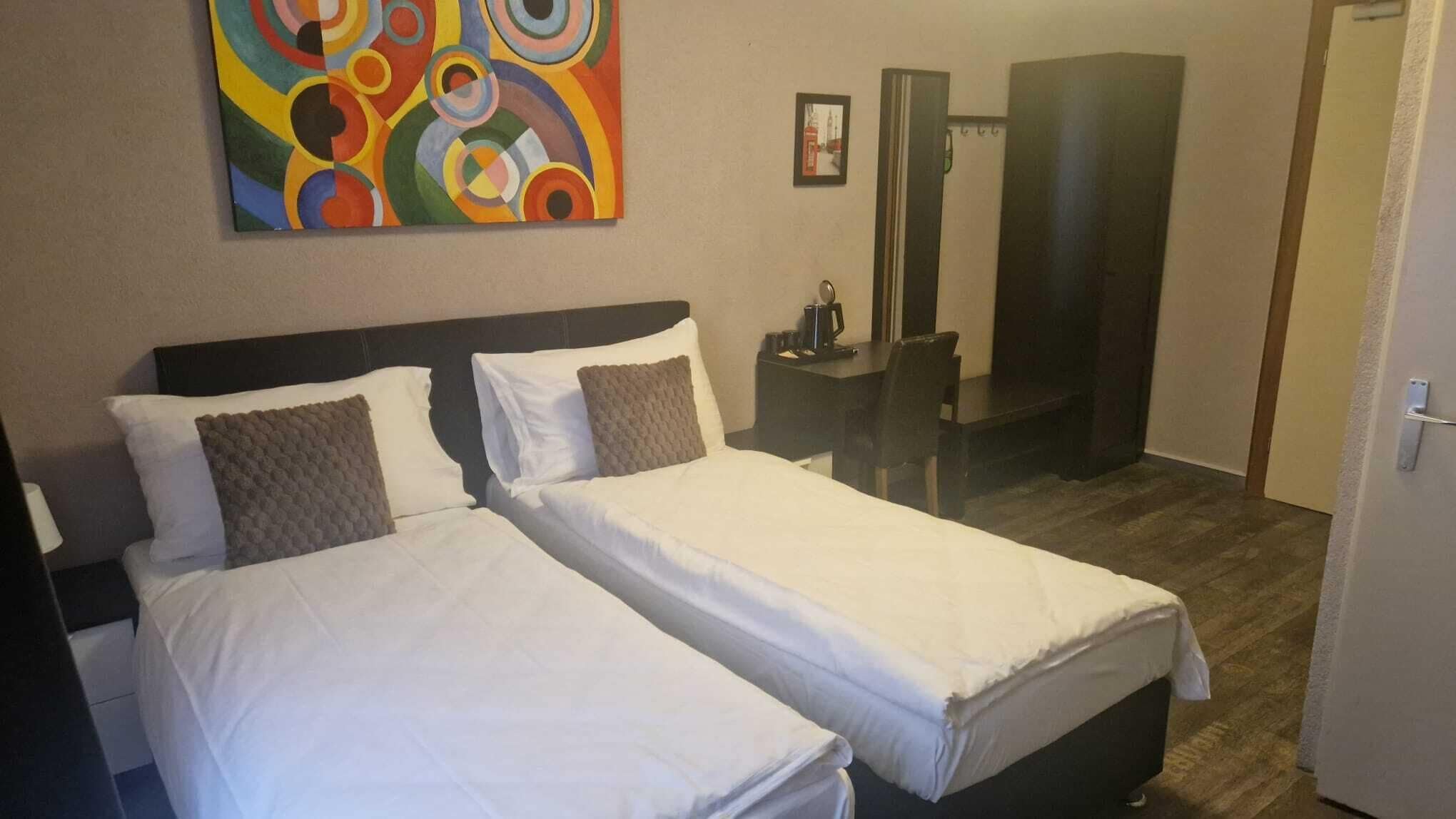 Ein Hotelzimmer mit zwei Einzelbetten, einem großen abstrakten Gemälde, einem kleinen Tisch mit einer Tasse und einem Spiegel.