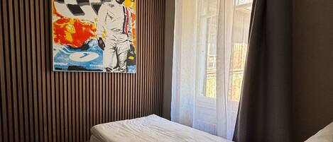 A room with a single bed, a wall-mounted painting, a window with curtains, and a ceiling light fixture.