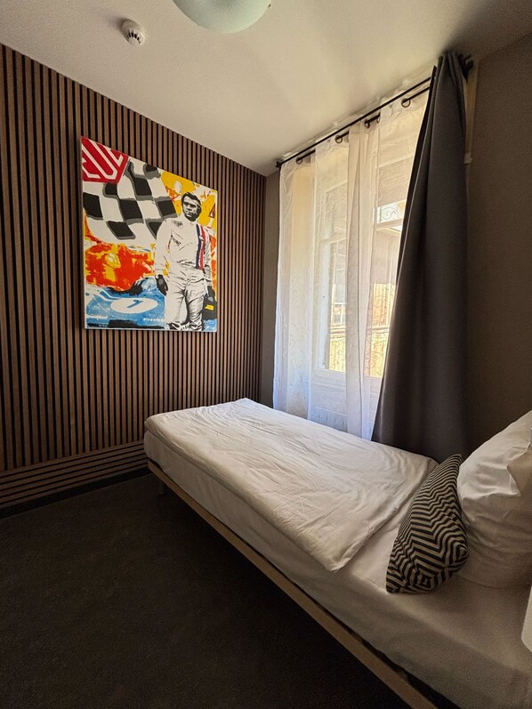 Una habitación con una cama individual, un cuadro colgado en la pared, una ventana con cortinas y una lámpara en el techo.