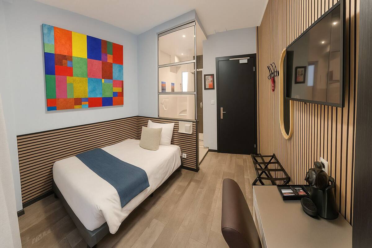 A modern hotel room with a bed, a desk, a chair, a colorful abstract painting, and a wardrobe.