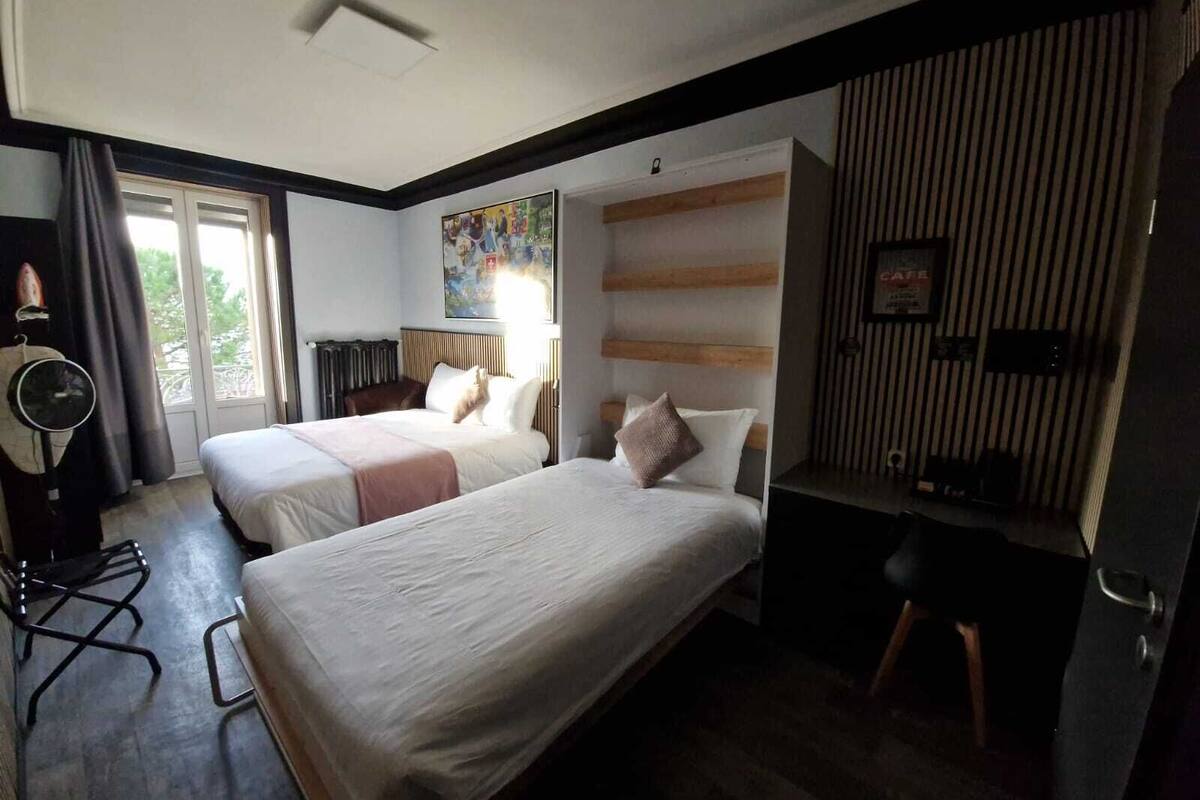 A hotel room with two beds, a desk, a chair, a fan, and a window with curtains.