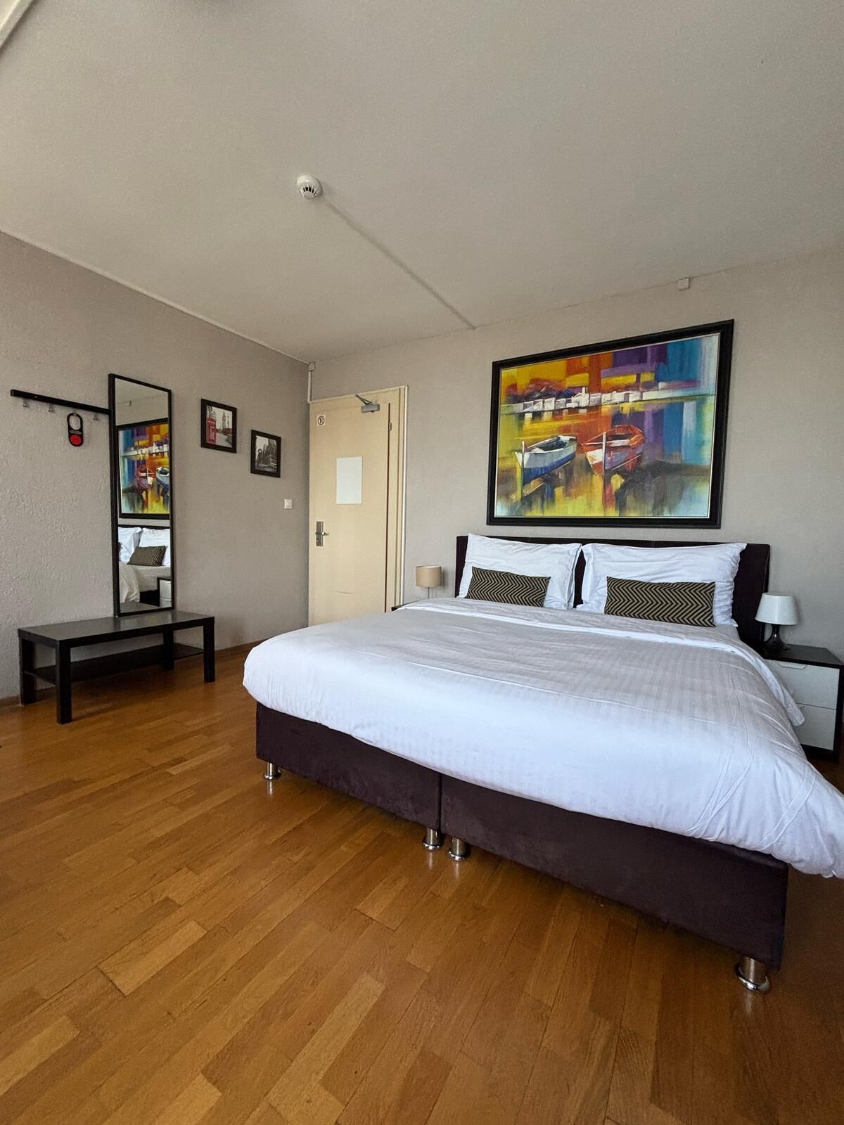 A modern bedroom with a large bed, a bedside table, a mirror, and a painting on the wall.