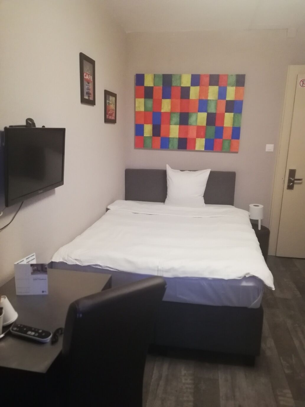 A hotel room with a bed, a TV mounted on the wall, a colorful artwork above the bed, and a desk with a chair.