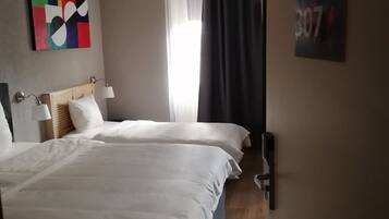 Una camera d'albergo con due letti, una lampada a soffitto, una finestra con tende e un quadro astratto dai colori vivaci appeso al muro.
