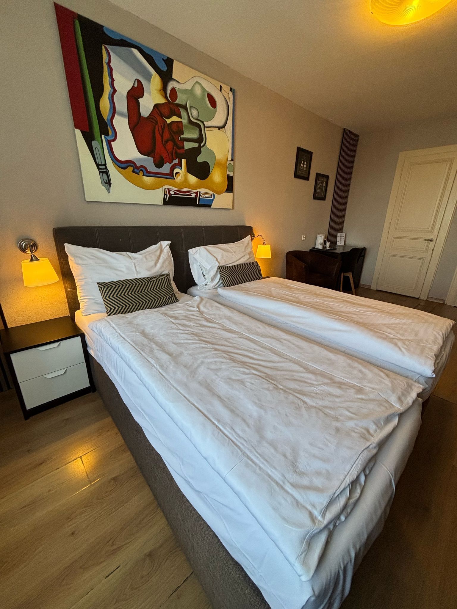 A double bed with white linens, a nightstand with a lamp, a painting on the wall, and a door in the background.