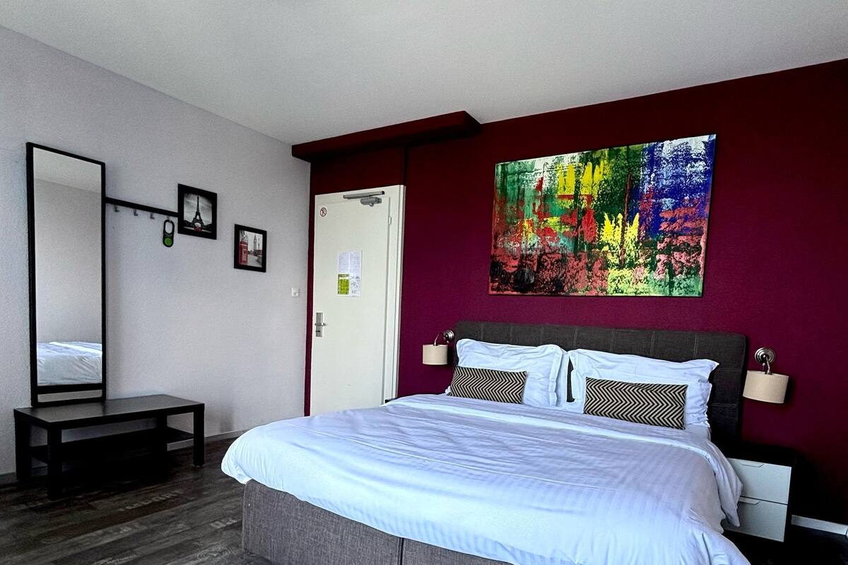A hotel room with a large bed, a bedside table, a mirror, and a colorful abstract painting on the wall.
