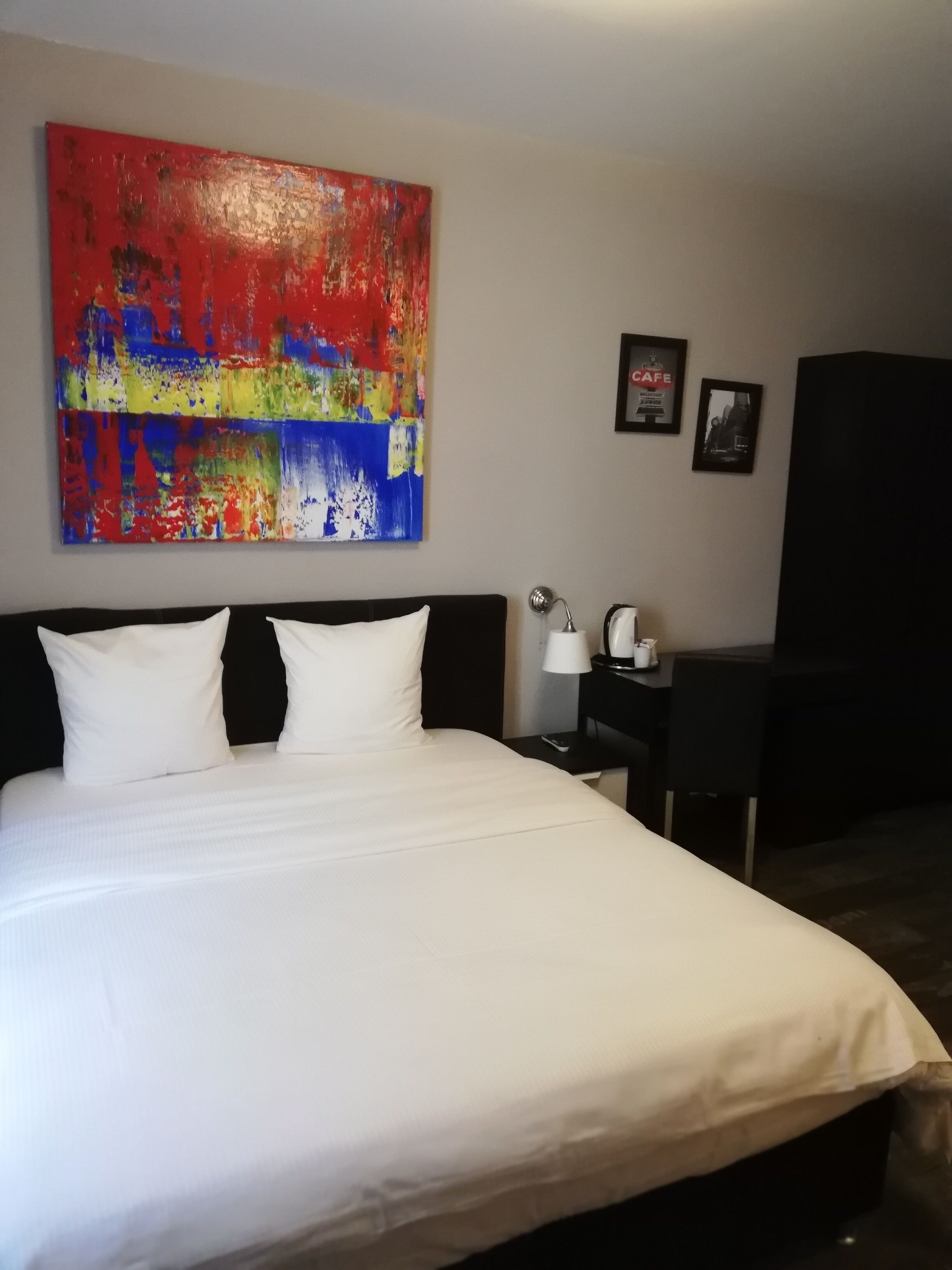 A hotel room with a large bed, a desk, a chair, a lamp, and a colorful abstract painting on the wall.