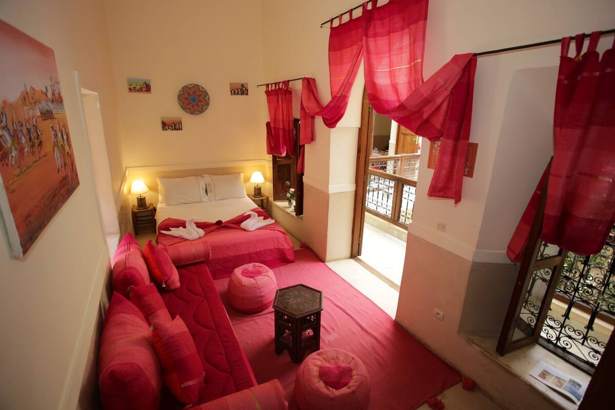 A room with a bed, two bedside tables, a window with red curtains, and a small wooden stool.
