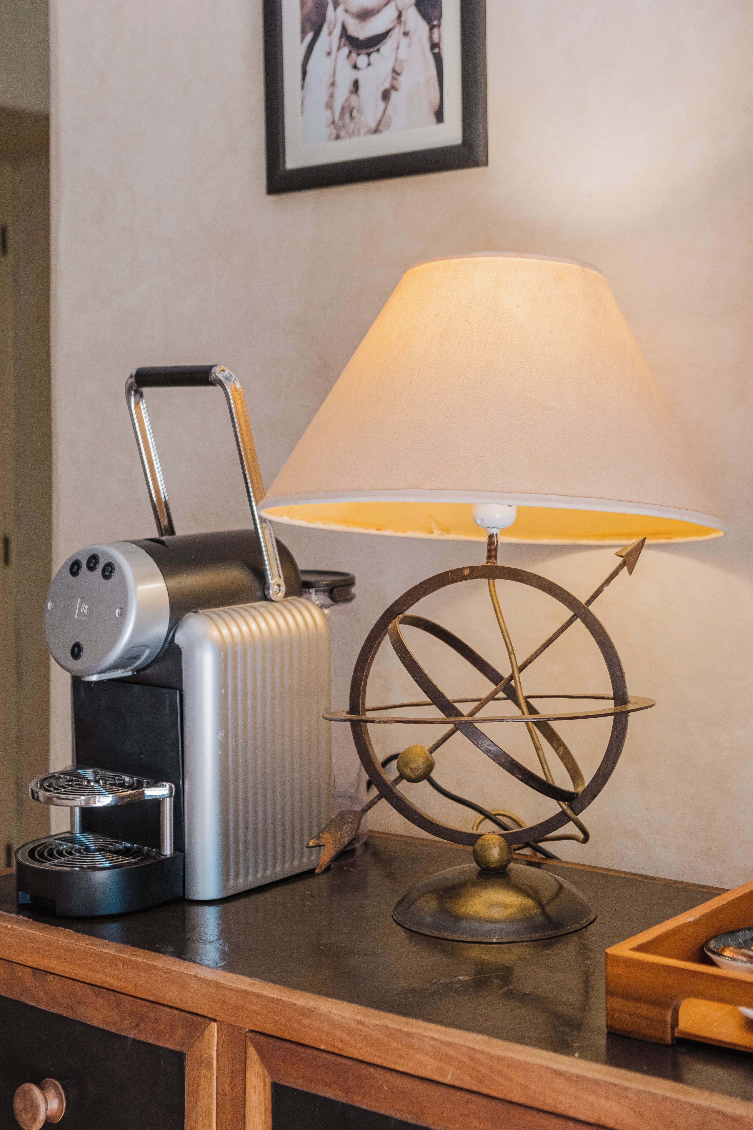 A kitchen with a coffee maker, a lamp with a metal base, and a framed picture on the wall.