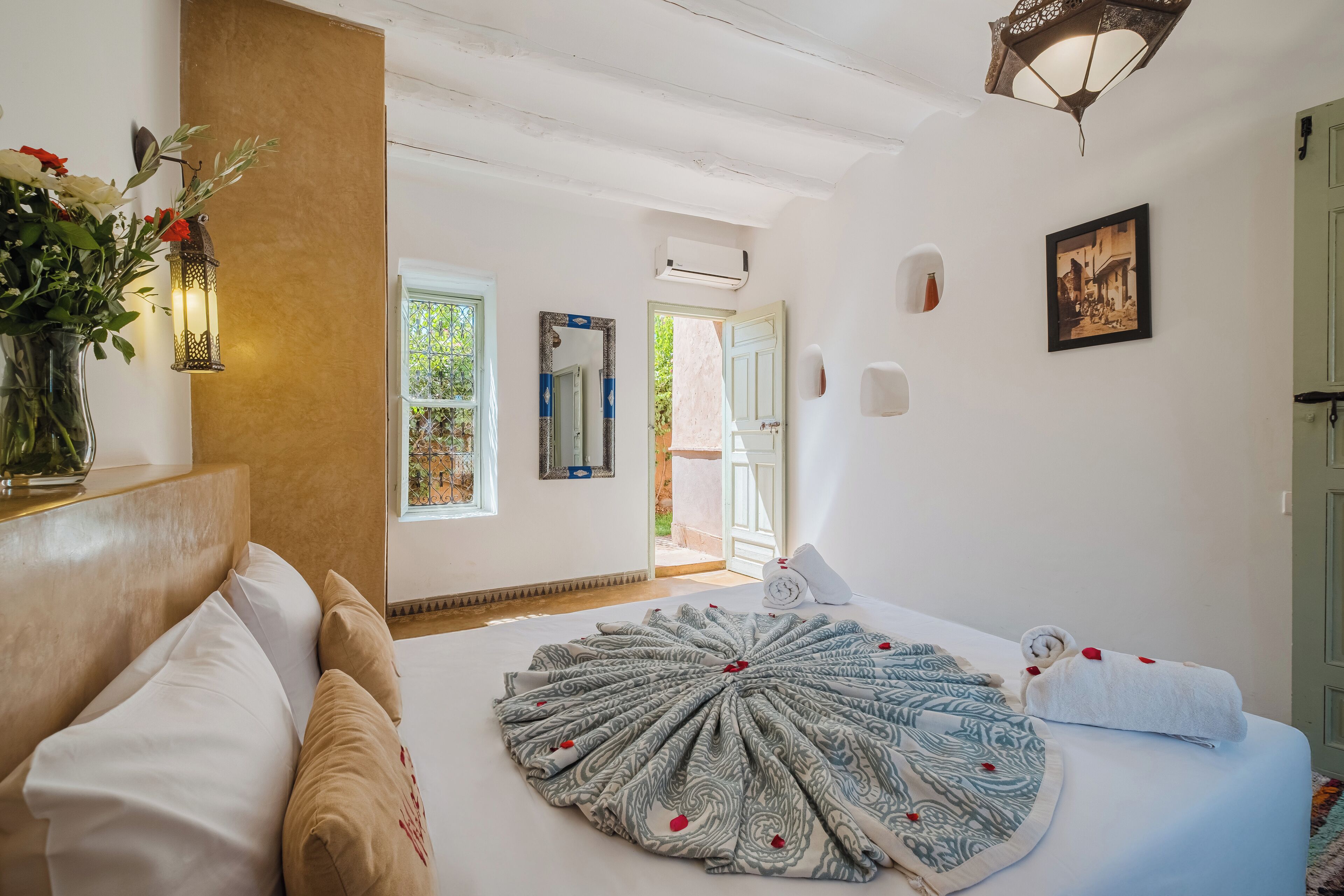 Une chambre dotée d’un grand lit, d’une table de chevet ornée d’un vase de fleurs, d’un climatiseur fixé au mur et d’une porte donnant sur un espace extérieur.