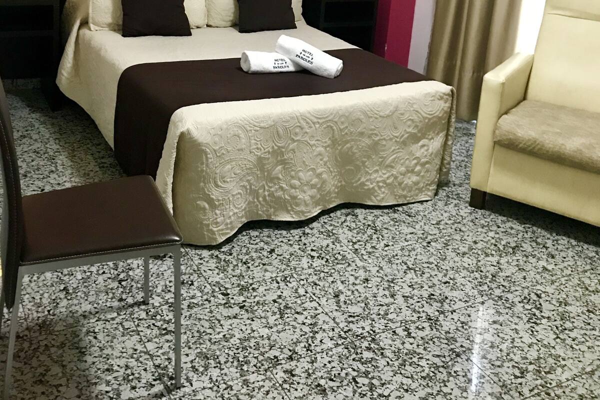 Une chambre d’hôtel comprenant un lit, une table de chevet, une chaise, une tête de lit, une œuvre d’art murale et un rideau.
