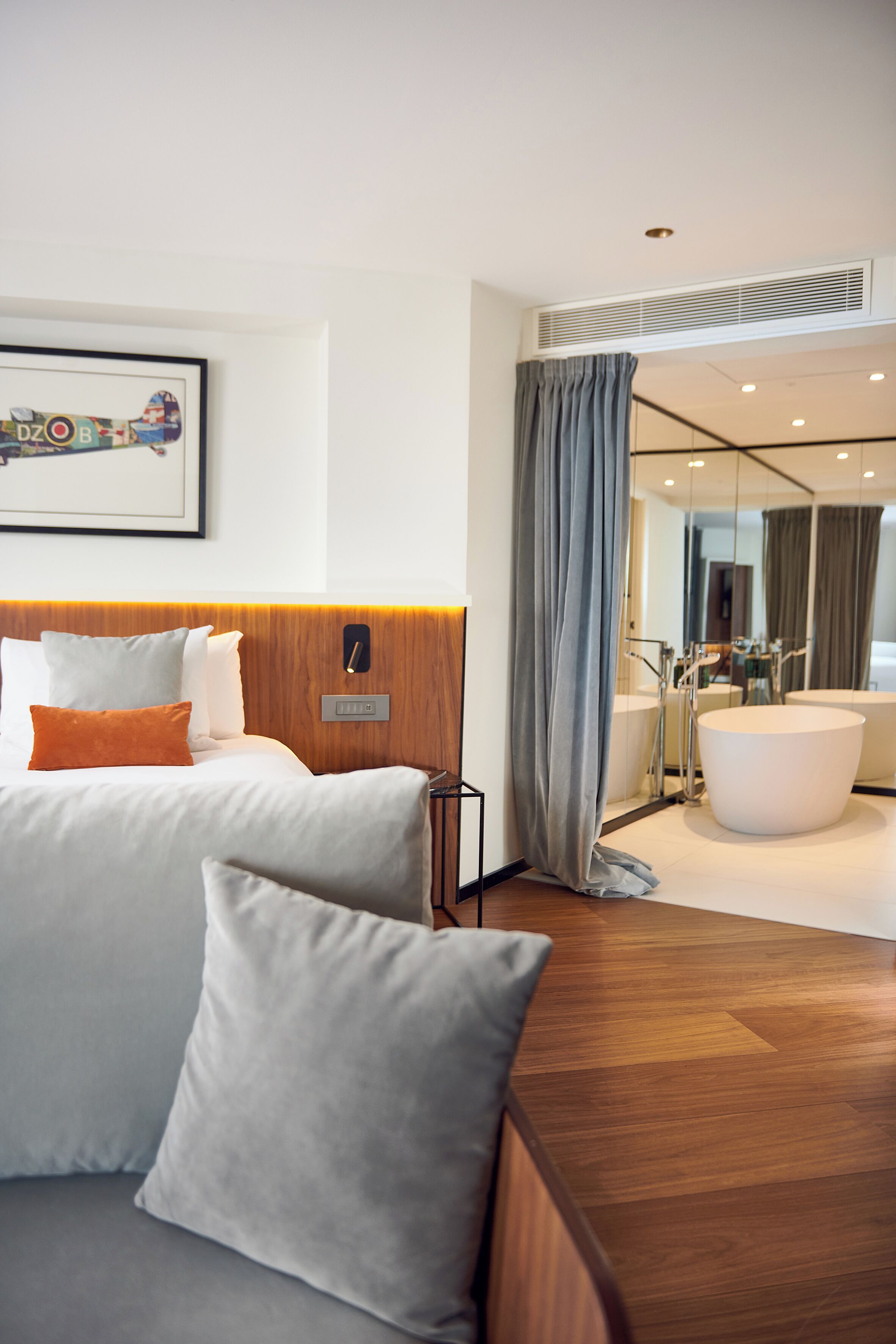 Una habitación de hotel moderna con tablero de cama de madera, una cama grande con almohadas blancas y anaranjadas, un sofá gris y una bañera grande.
