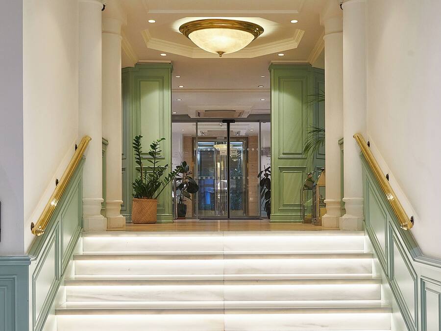 Una gran escalera conduce a un vestíbulo con entrada de puerta de cristal, flanqueado por columnas verdes y un candelabro.