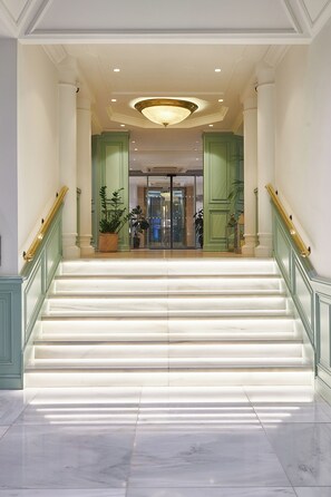 A grand staircase leading to a lobby with a glass door entrance, flanked by green columns and a chandelier. - The Level at the Melia White House (London)