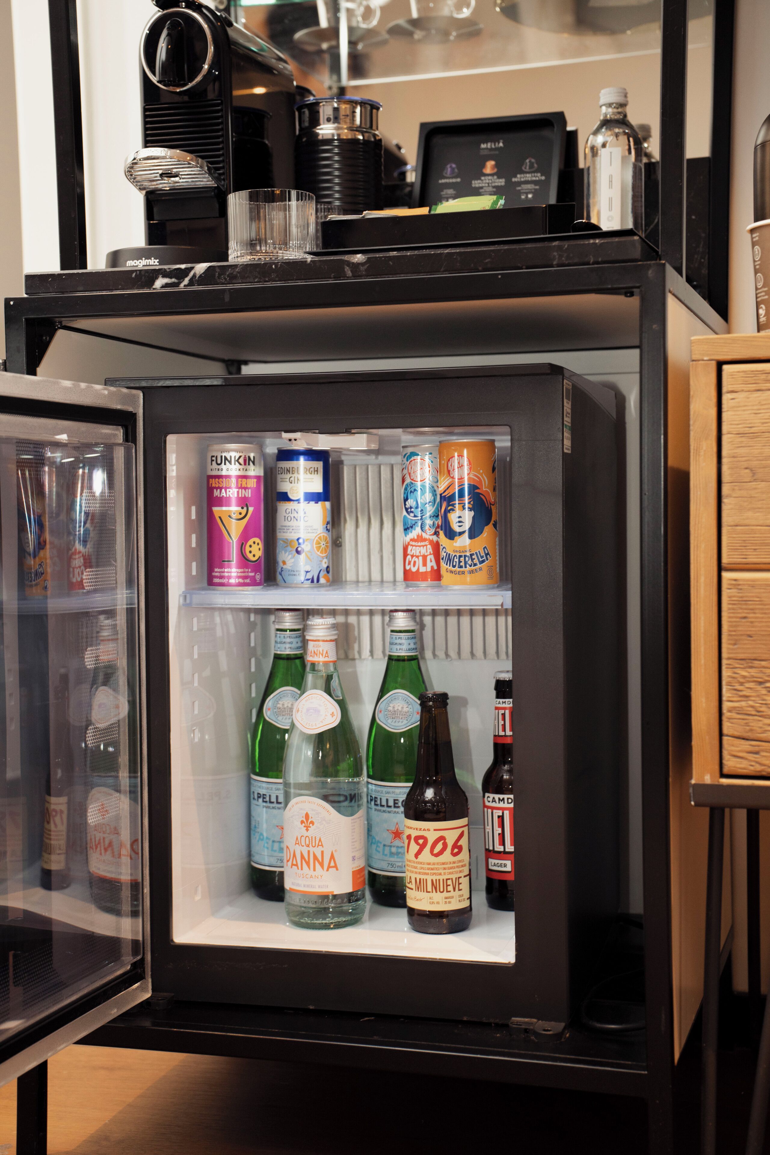 Un minibár con una cafetera, un certificado enmarcado y diversas bebidas, incluyendo Martini, ginebra, cola y cerveza.