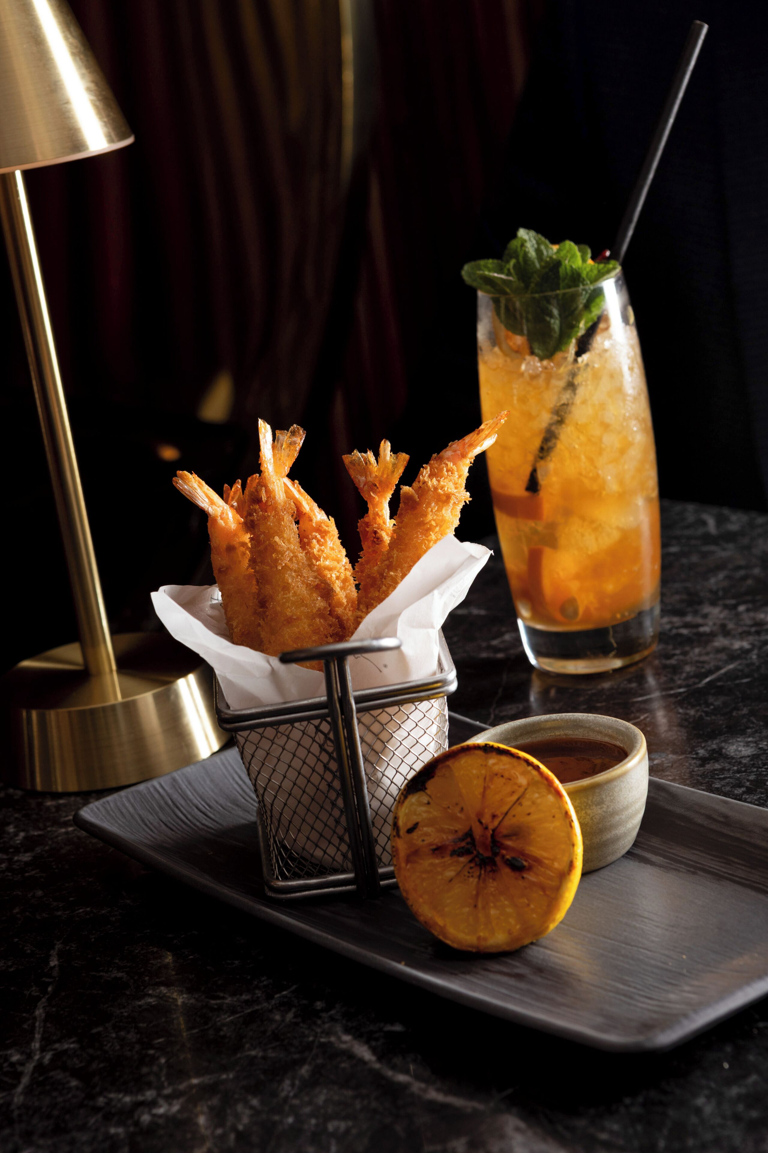 A tray with fried shrimp, a glass of iced drink with a straw, and a slice of orange on a dark marble surface.